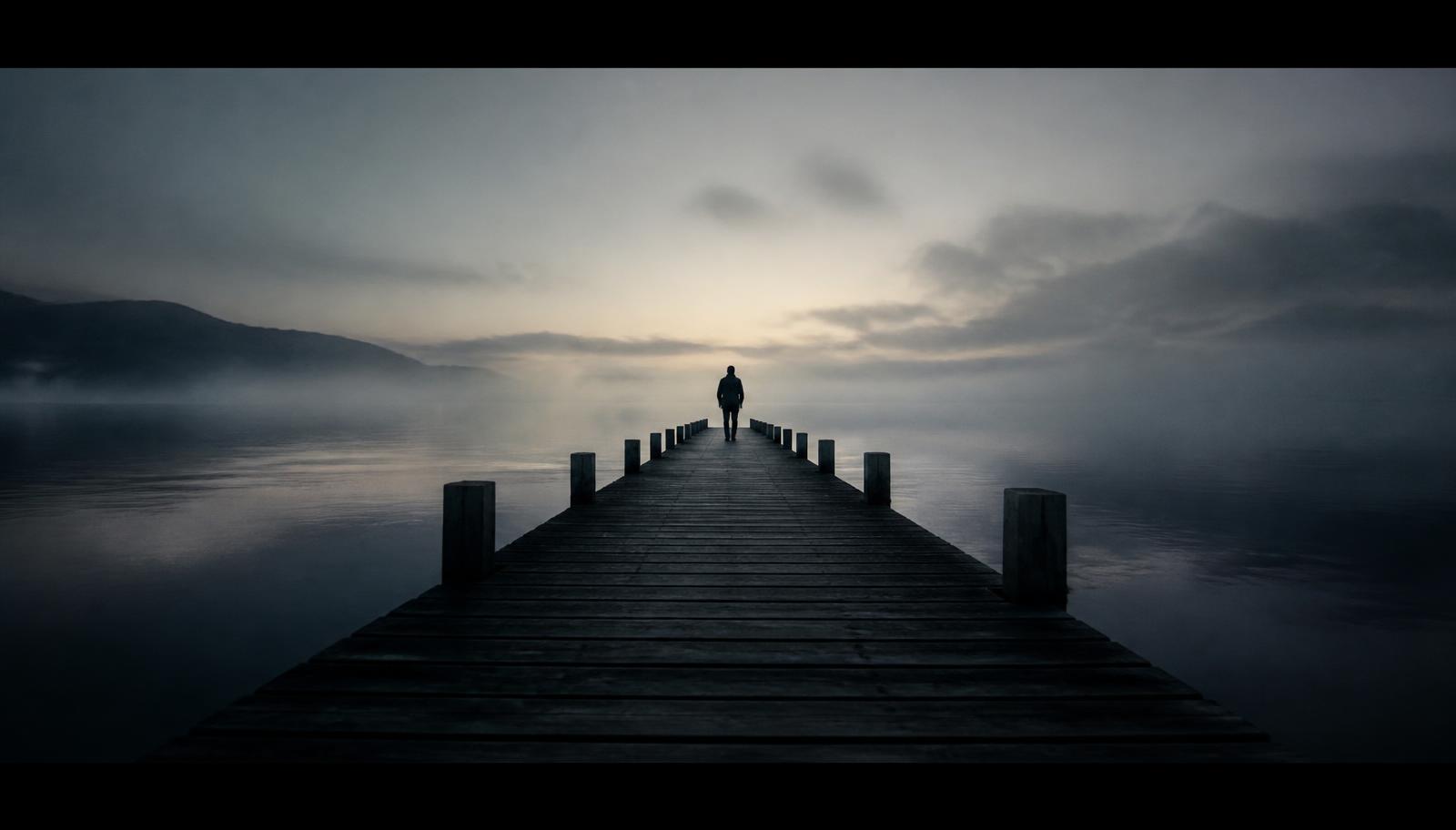A lone figure walking a wooden pier into ocean fog at first light